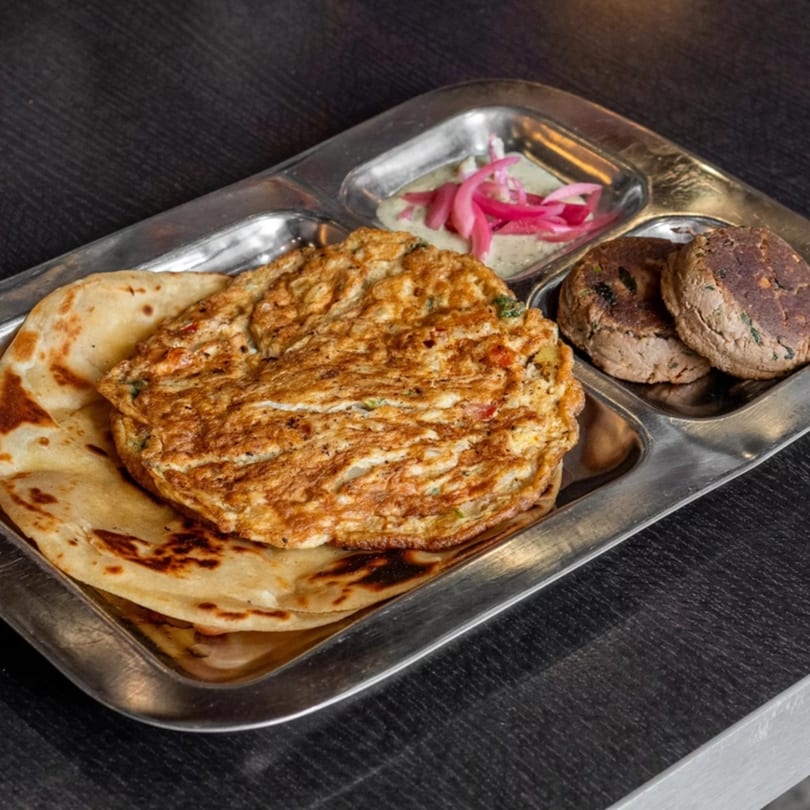 A metal tray with a Mirchi Cafe omelet, paratha, two brown patties, and a small dish of pickled onions.