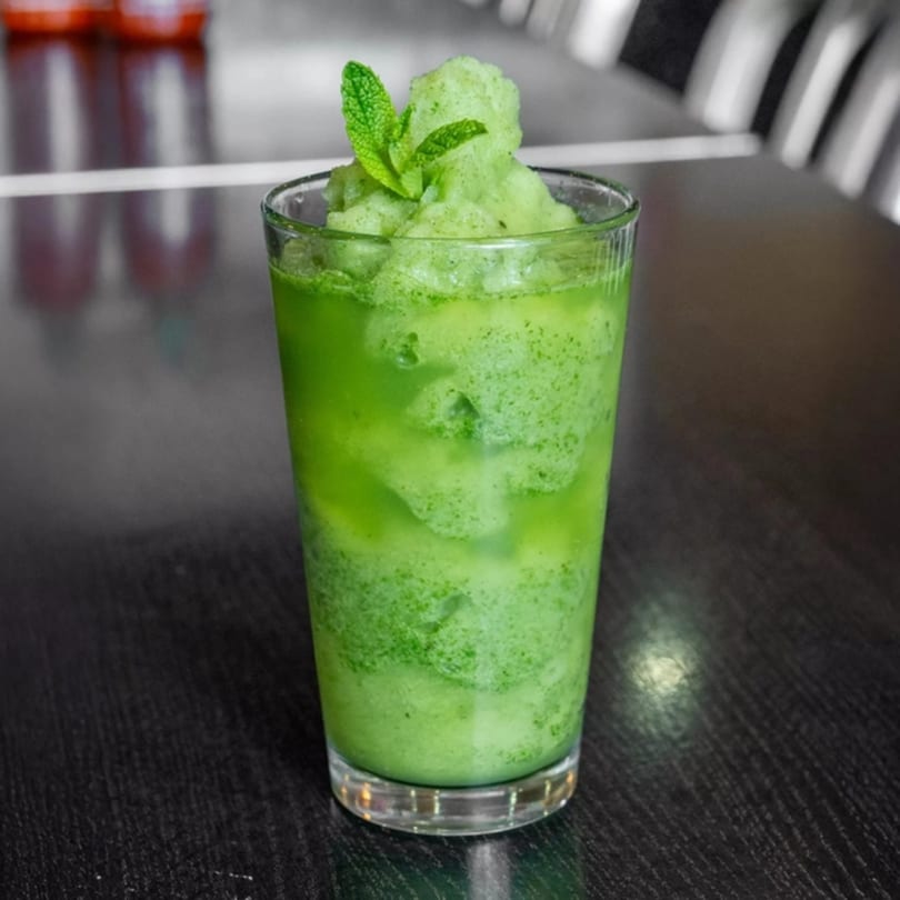 A tall glass of vibrant green mint slushie, topped with fresh mint leaves, on a dark table.