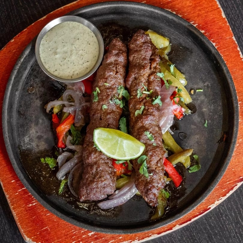 Two Mirchi Cafe seekh kebabs with bell peppers, onions, cilantro, lime, and green sauce on a black plate.