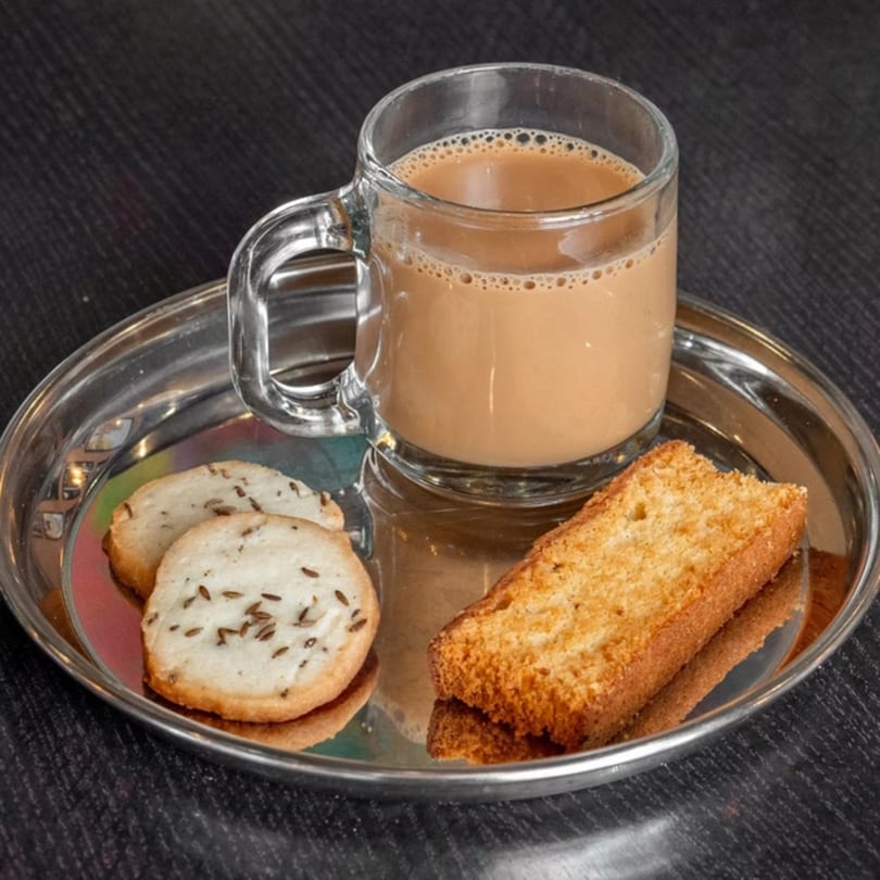 A glass mug of chai tea, two cumin cookies, and a rusk on a silver tray.