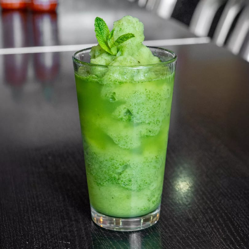 A vibrant green mint slushie in a clear glass, garnished with fresh mint leaves on a dark table.